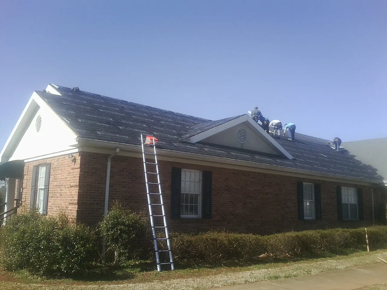 Crew tearing shingles off a brick ranch roof with a ladder set — Emergency Roof Repair work by our crew in Batchelor