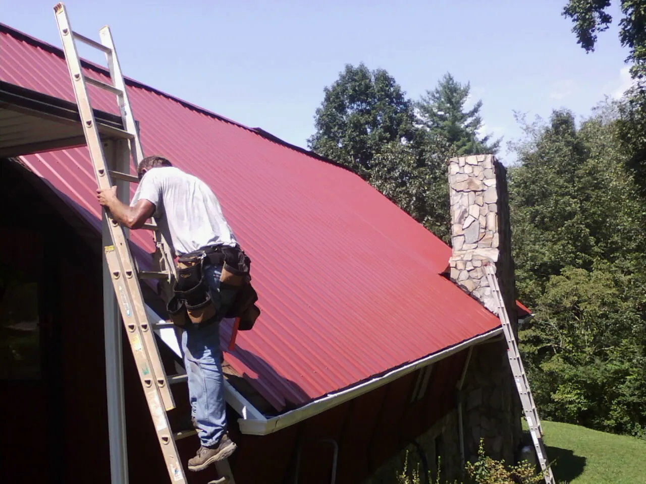 Roofer on a ladder beside a red metal roof and stone chimney — Soffit & Fascia Repair by Roofing Brothers in Batchelor